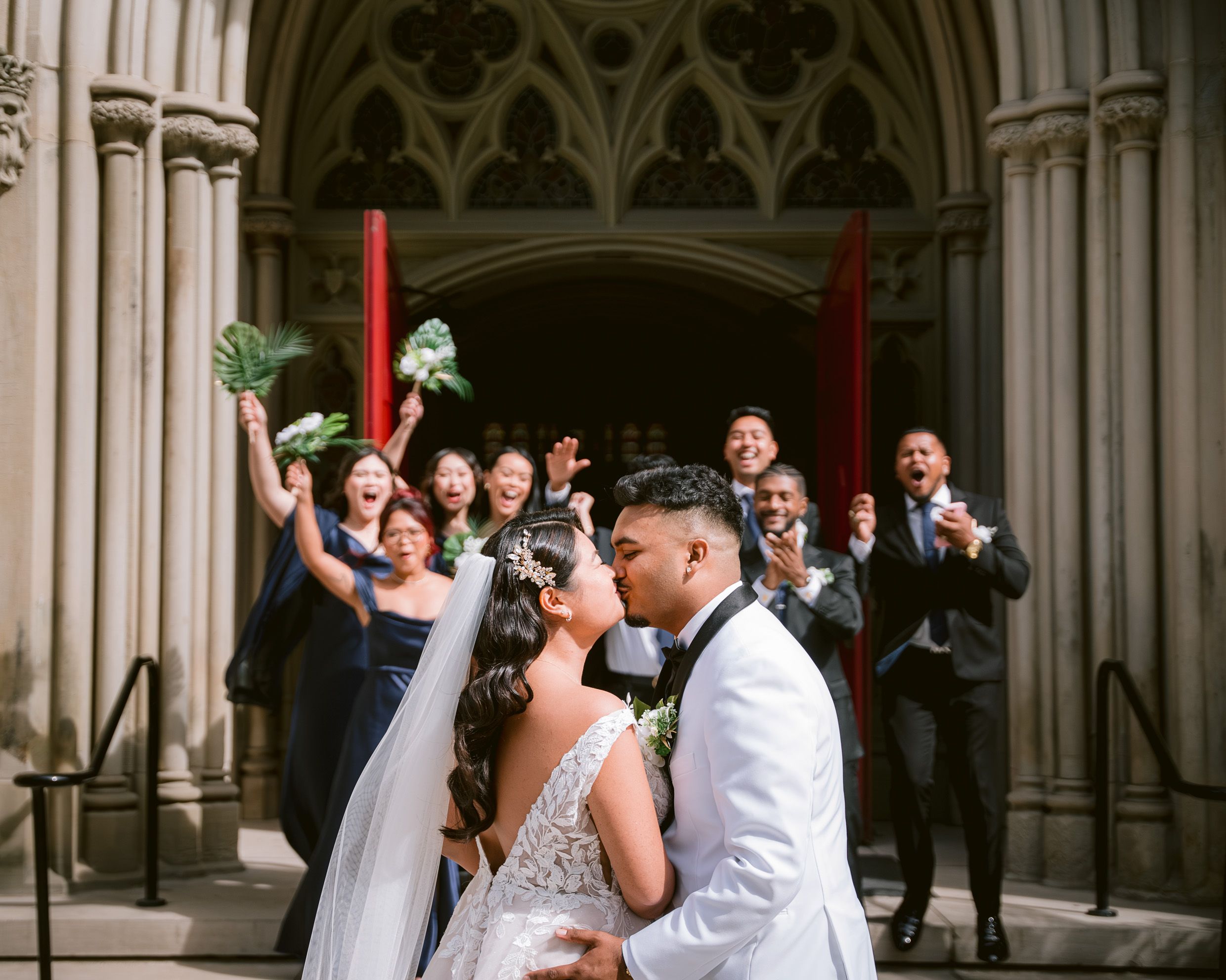 Wedding celebration with group at church entrance while newlyweds share first kiss.