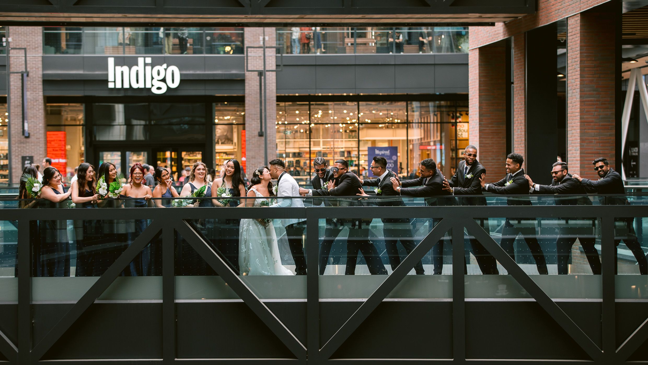 Crowd of people gathered on a pedestrian bridge outside an Indigo storefront during dusk.