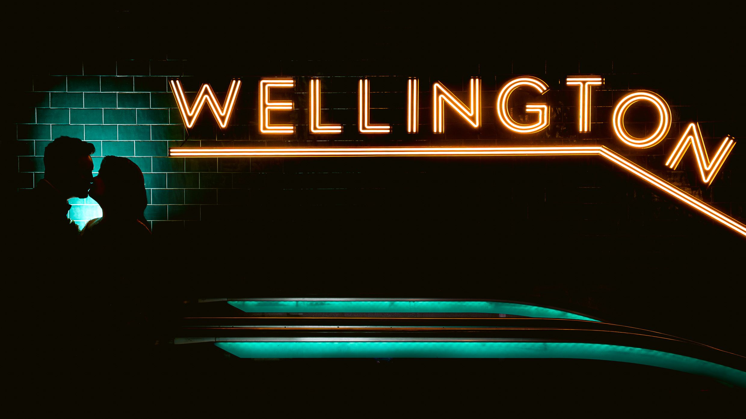 Illuminated Wellington sign glows in turquoise against dark urban wall.