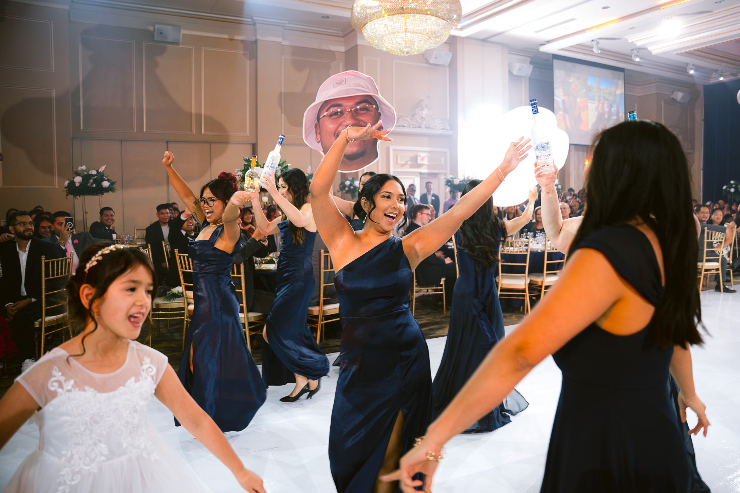 Wedding reception guests in navy blue dresses dancing and celebrating on a ballroom dance floor under chandeliers.