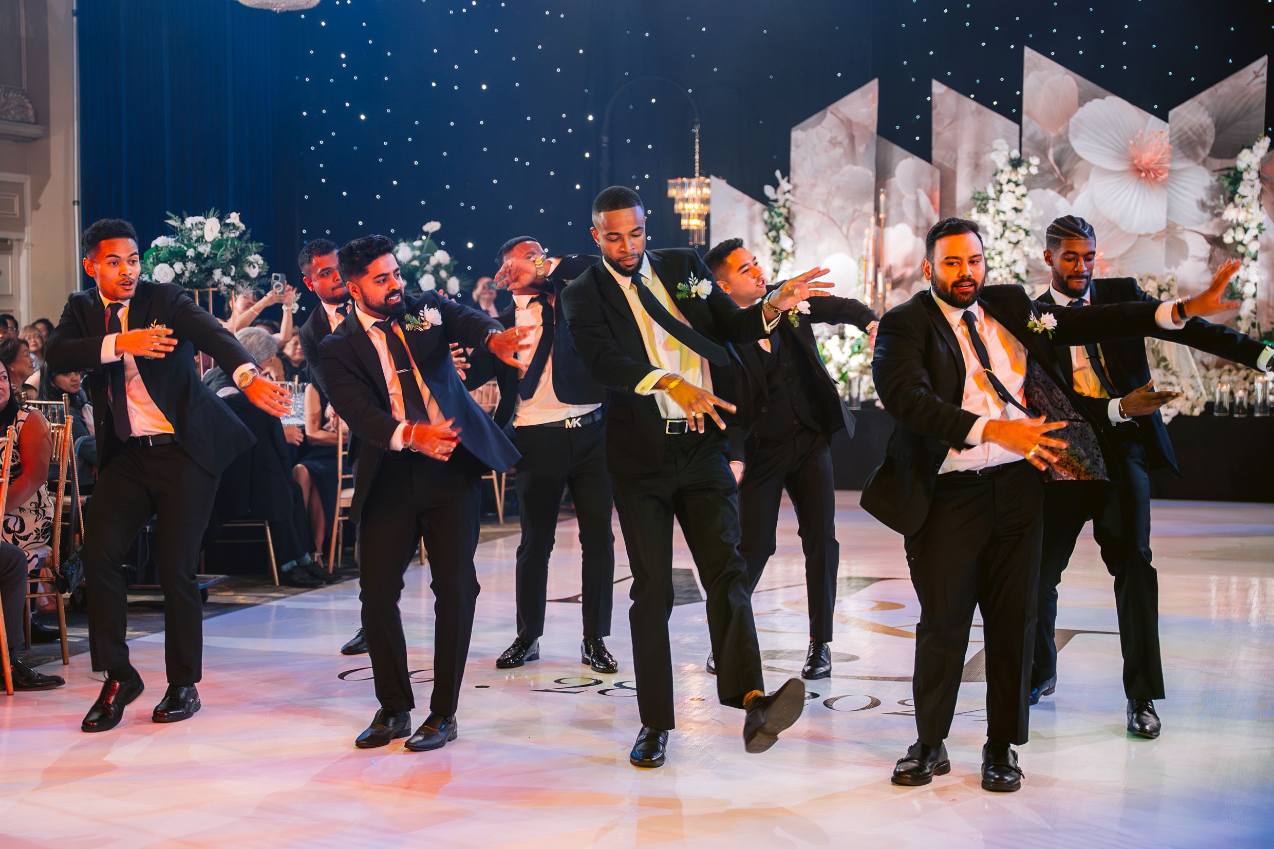 Wedding party performs choreographed dance routine on illuminated floor with festive backdrop.