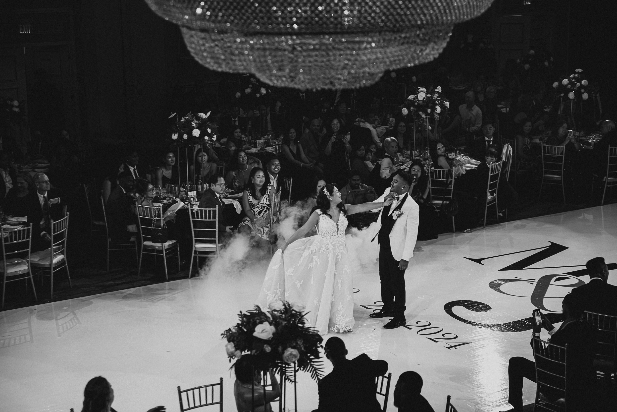 Black and white photo of a bride and groom sharing their first dance under a crystal chandelier at an elegant reception.