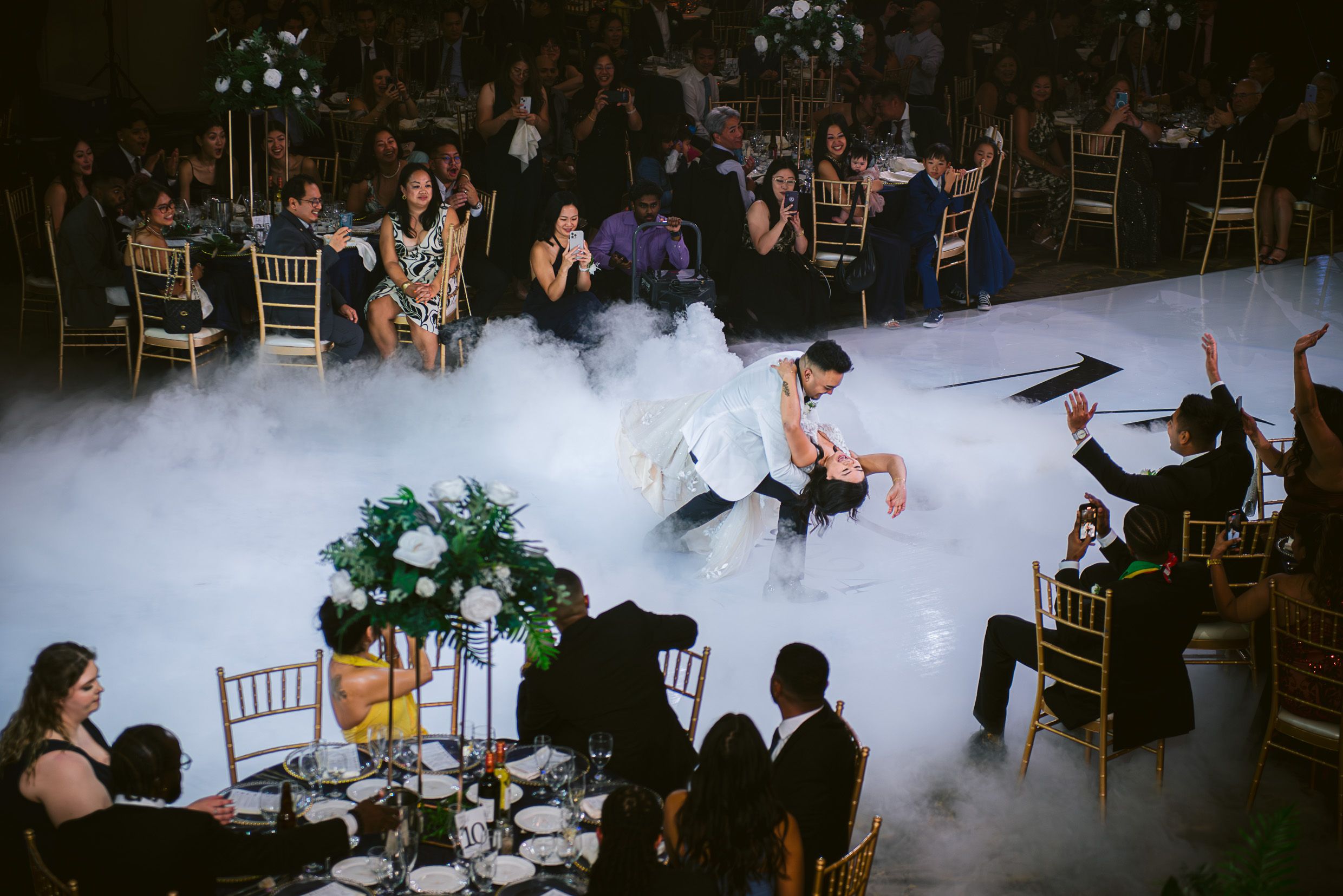 A couple dancing on a fog-filled dance floor during a wedding reception as guests watch from surrounding tables.