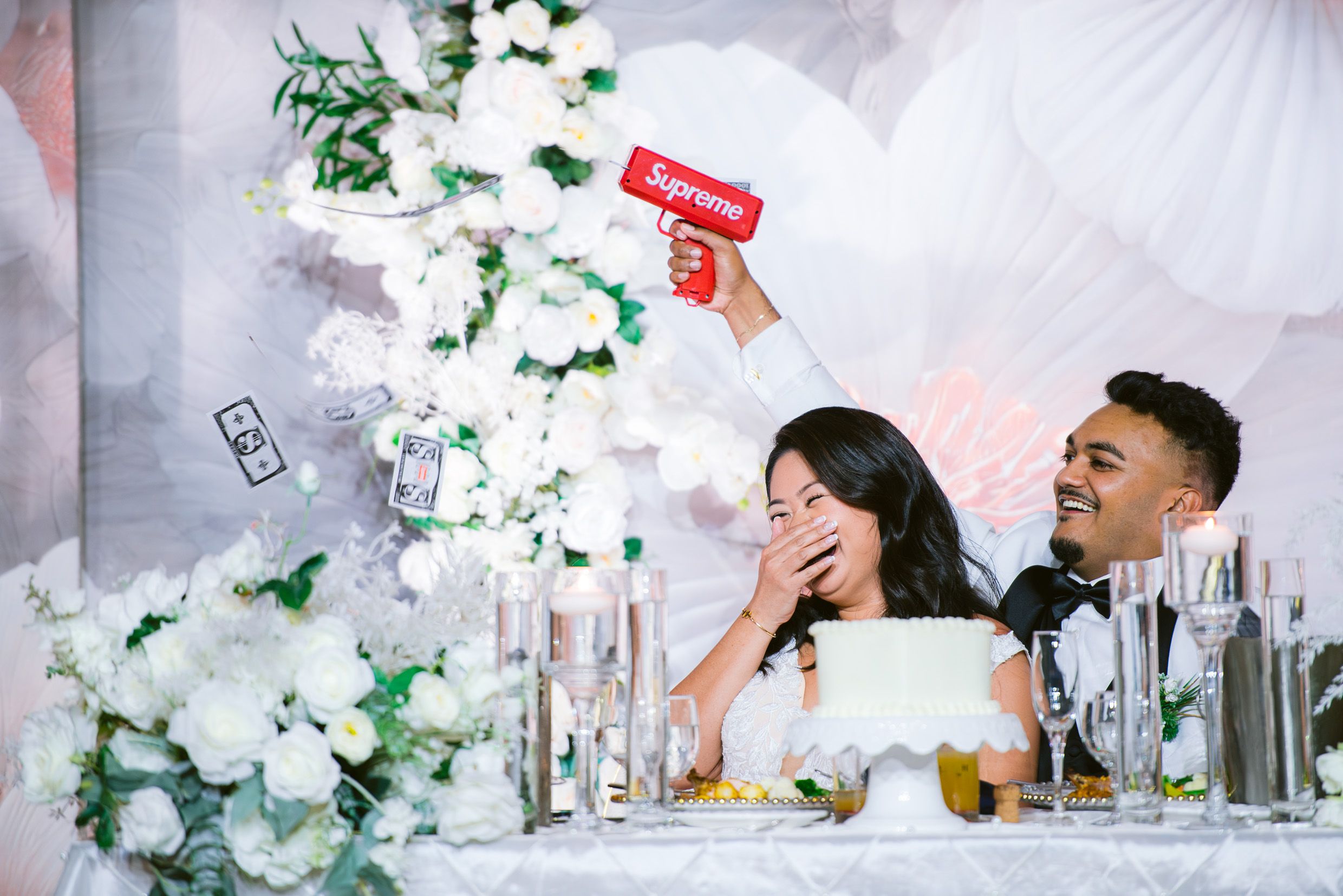 An intimate wedding reception scene with laughter shared at a white-decorated table with floral arrangements.