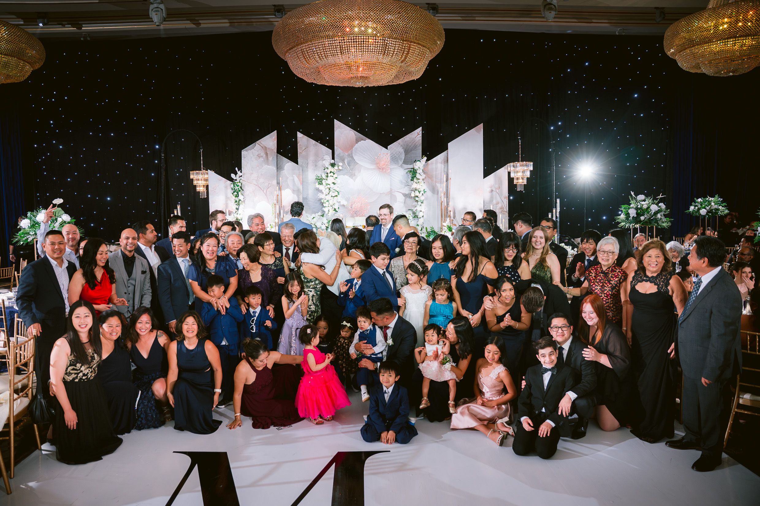 A large wedding party poses together on a dance floor with geometric decorations and hanging lights during a reception.