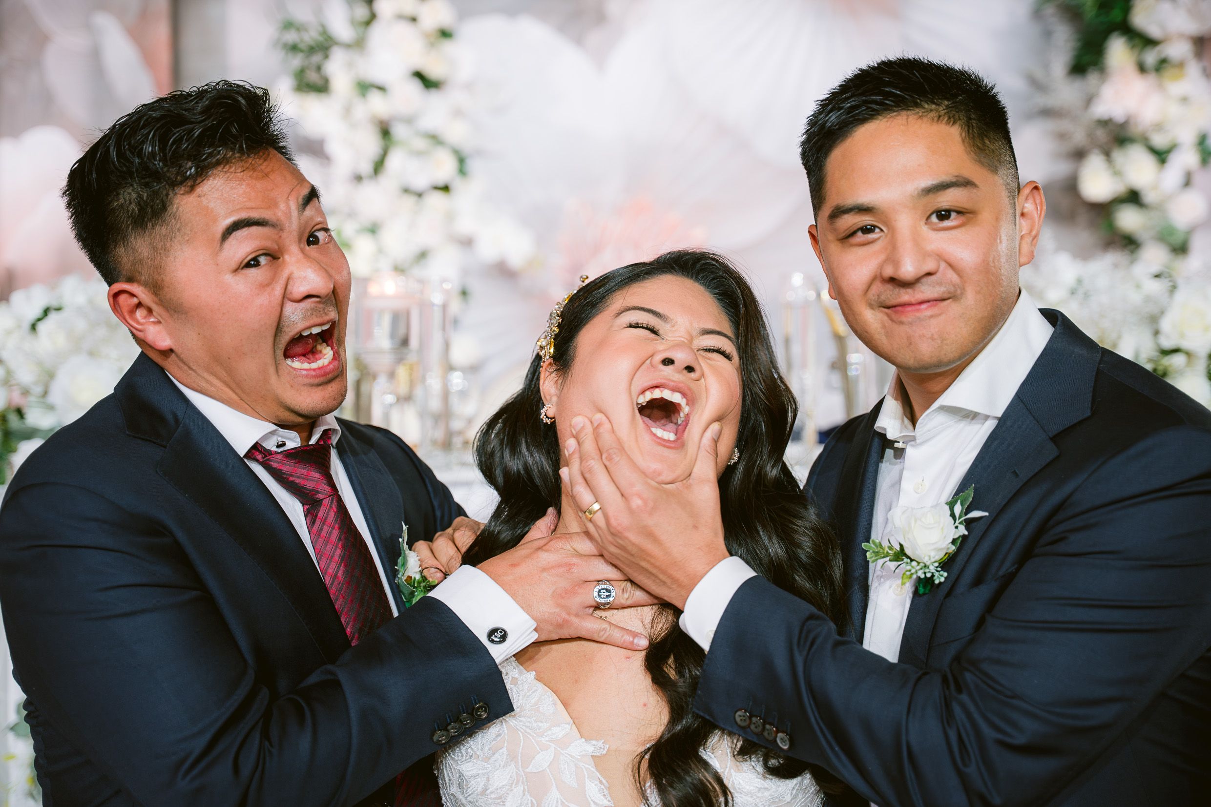Three people share a candid joyful moment together during a wedding celebration with white floral decorations in background.