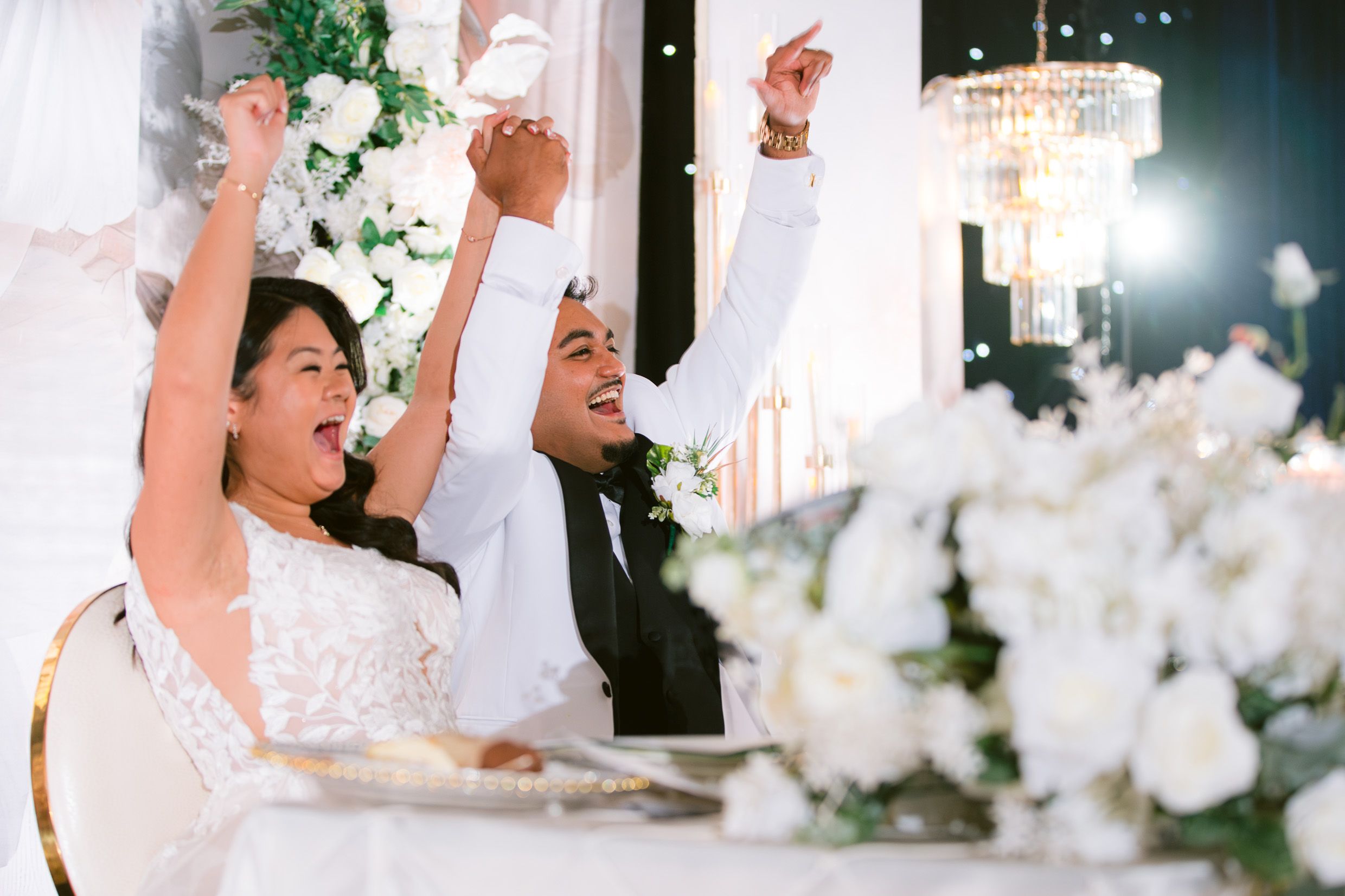A celebratory moment at an elegant wedding reception with white floral decorations and a crystal chandelier overhead.