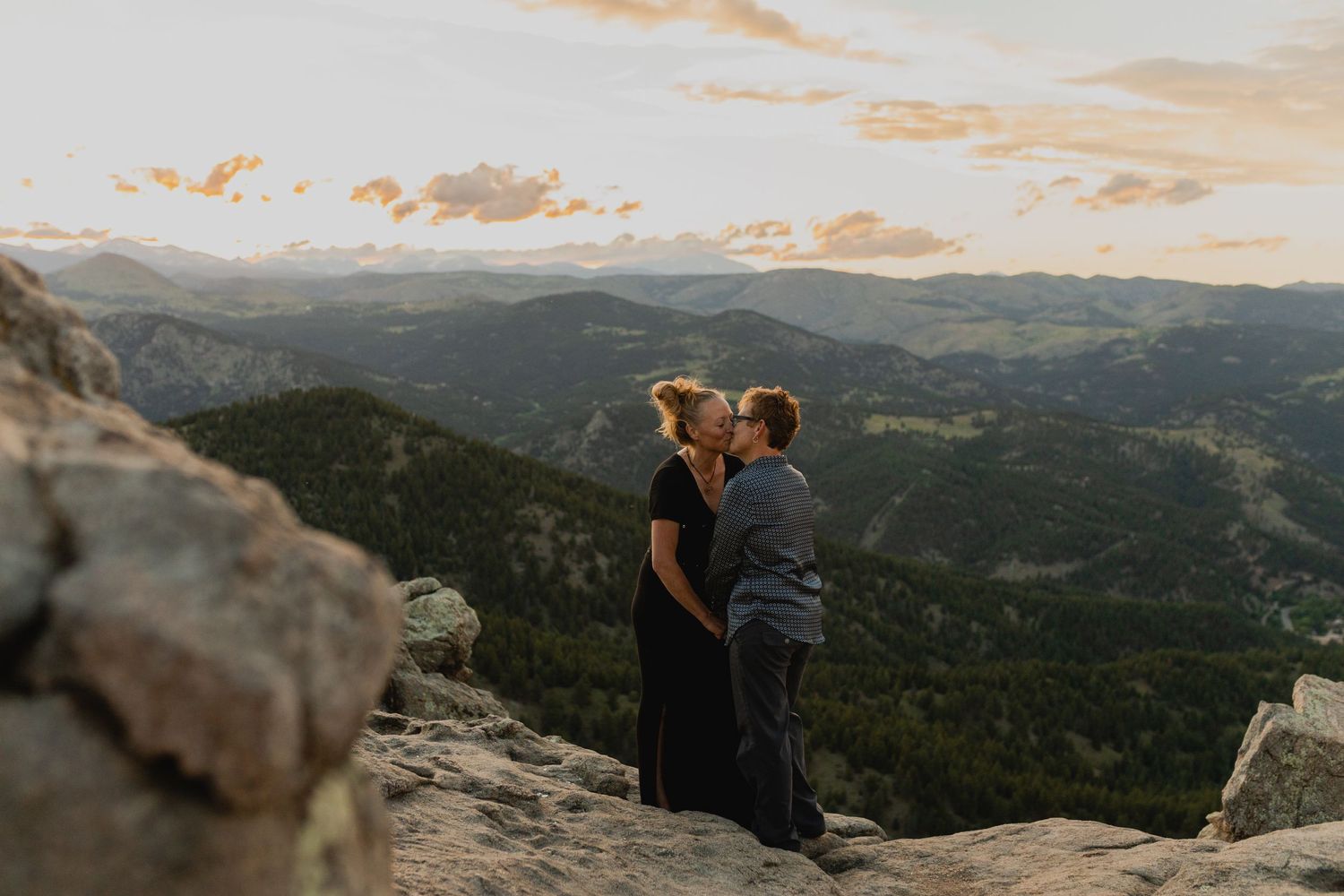 Two figures stand at sunset on a mountain overlook with panoramic views of rolling green hills and distant peaks.