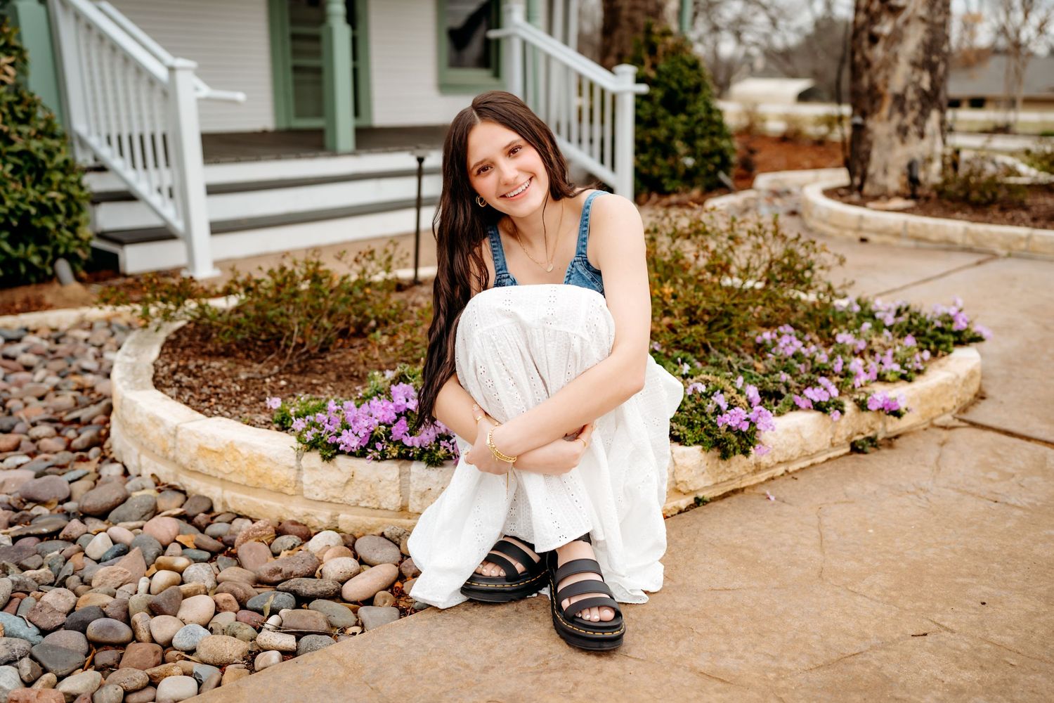 Person sitting on a stone pathway wearing a white dress and sandals next to a garden with purple flowers.
