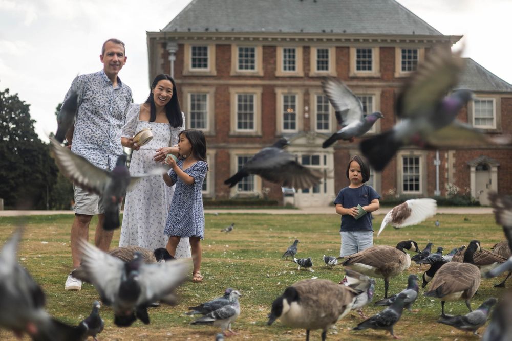 Family of 4 in front of Forty Hall in Enfield. The family are feeding the pigeons and there are pigeons flying around them.