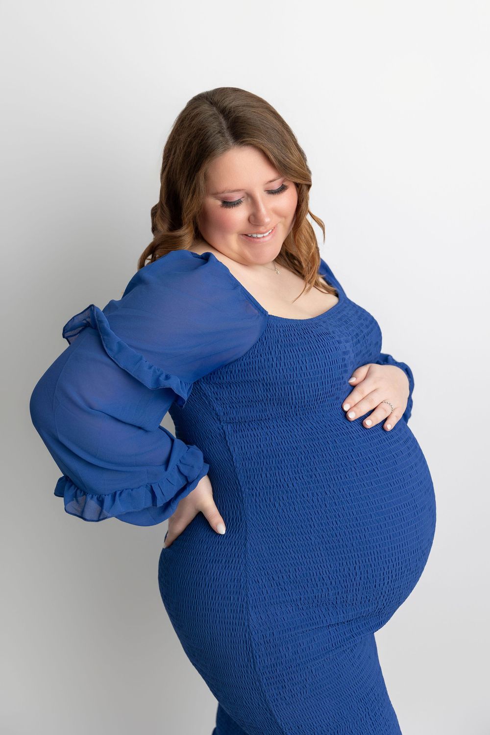 Expectant mother wearing royal blue ruched dress with sheer puff sleeves poses against white background in maternity photo.