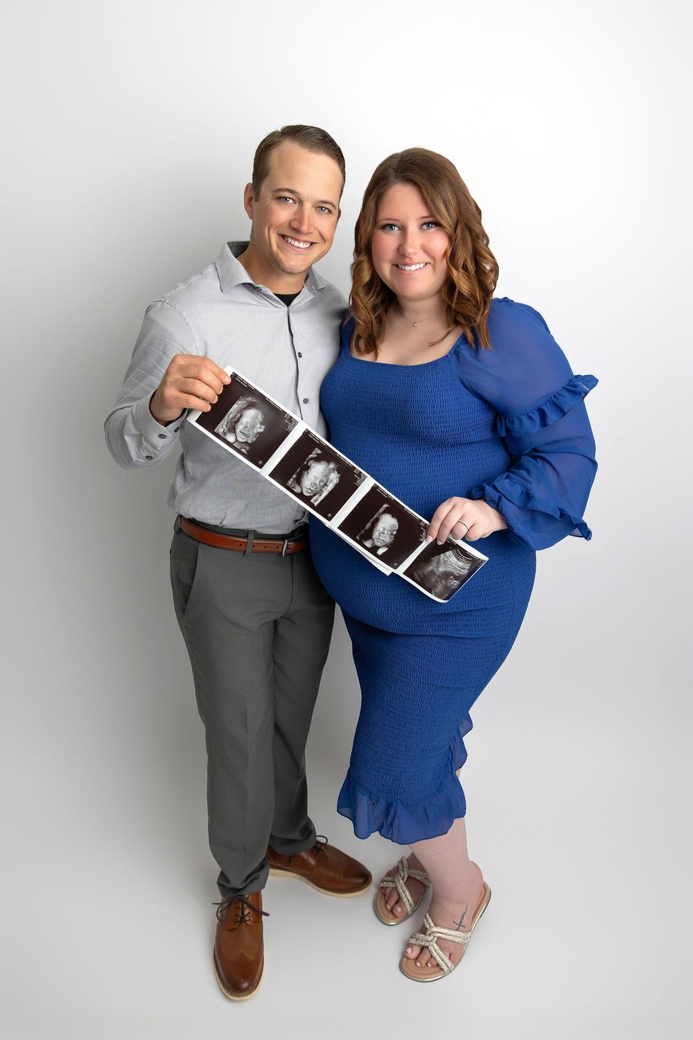 Expectant couple holding ultrasound images while standing against white backdrop, wearing grey and blue outfits.
