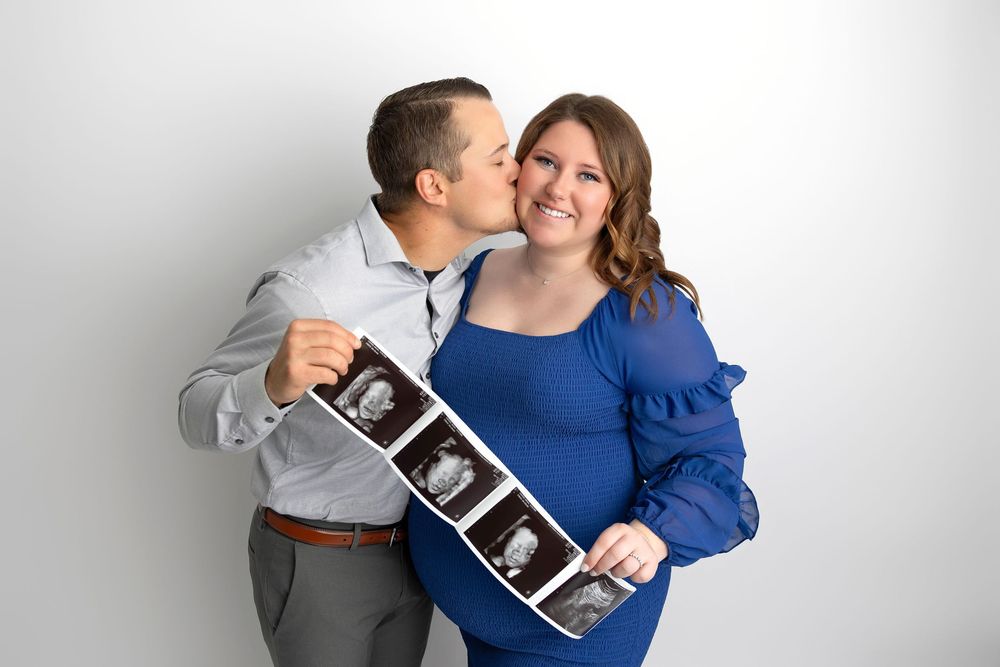 Couple in grey shirt and blue blouse share tender moment while holding ultrasound photos against white wall.