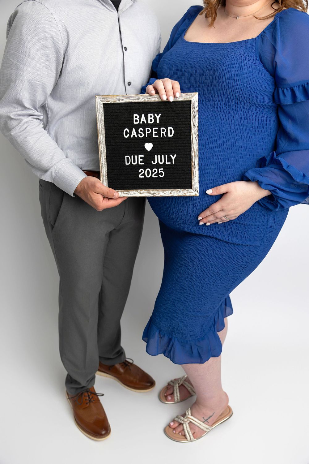 Expectant couple holds letterboard announcing Baby Caspero due July 2025 while wearing blue dress and grey outfit.