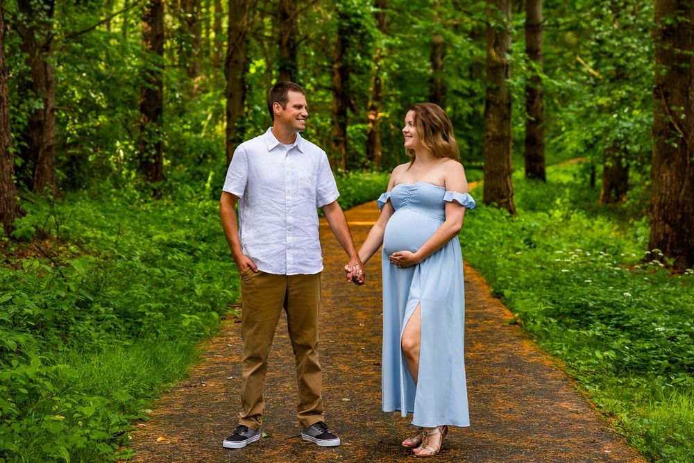 Expectant couple holds hands while walking down a lush forest path surrounded by vibrant greenery on a summer day.