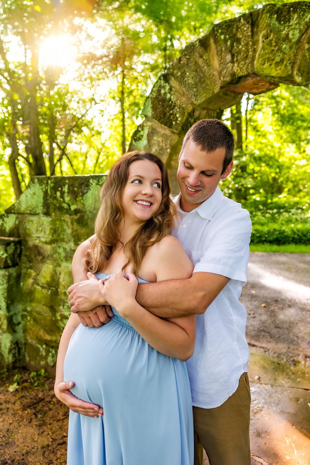 A couple embraces while standing in a wooded area during golden hour sunlight.