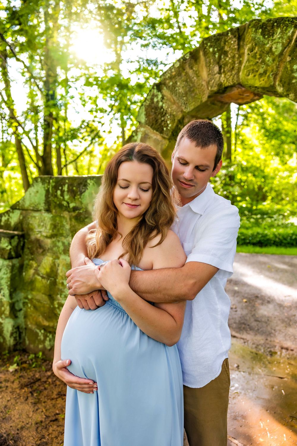 A romantic maternity photoshoot shows a couple embracing under a stone archway in a lush green garden setting.