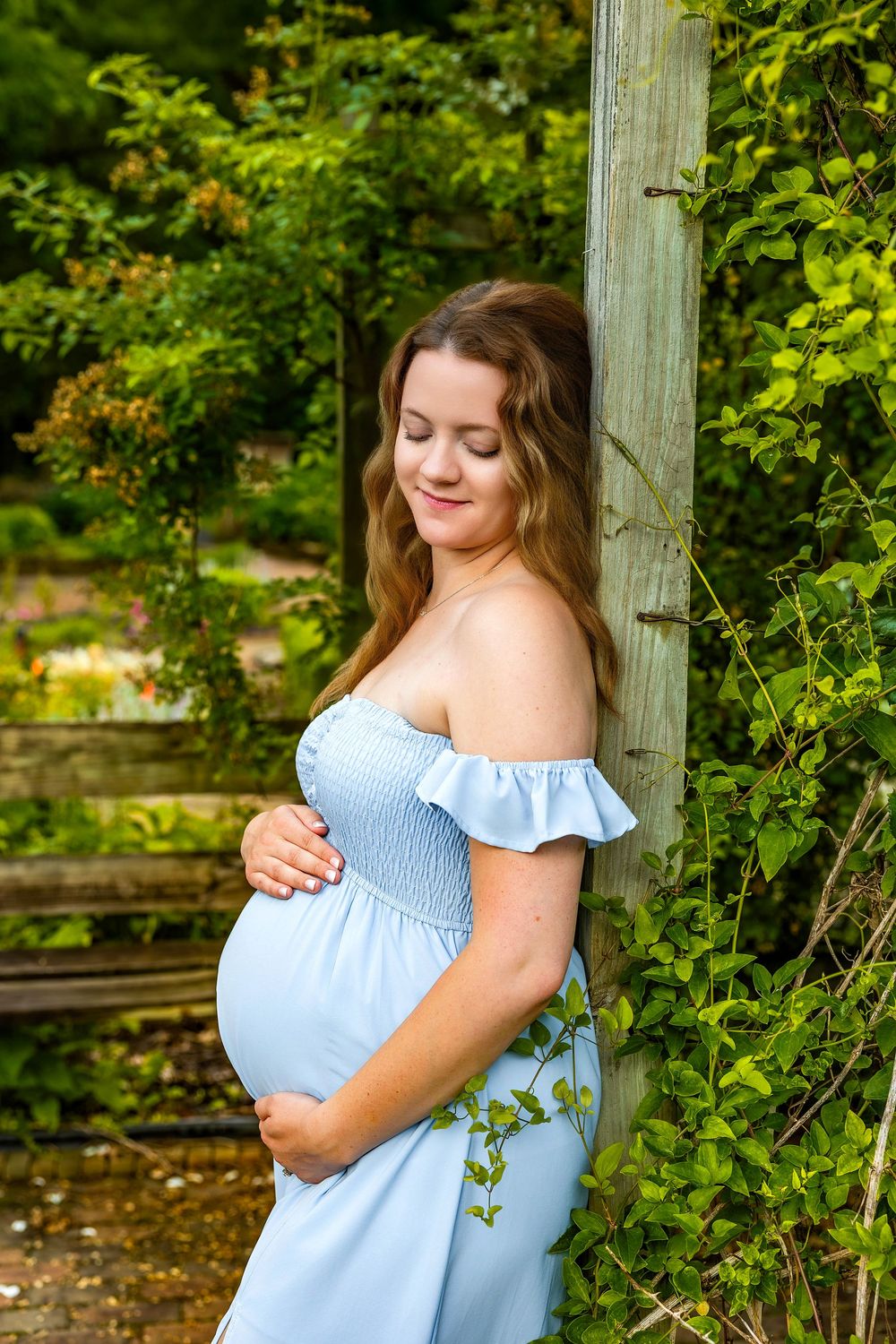A person in a light blue off-shoulder dress poses outdoors in a garden setting while cradling their pregnant belly.