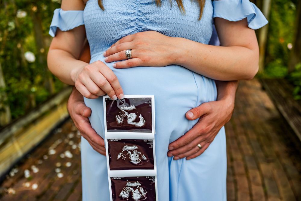 Someone in a light blue dress holds ultrasound images against their pregnant belly in an outdoor setting.