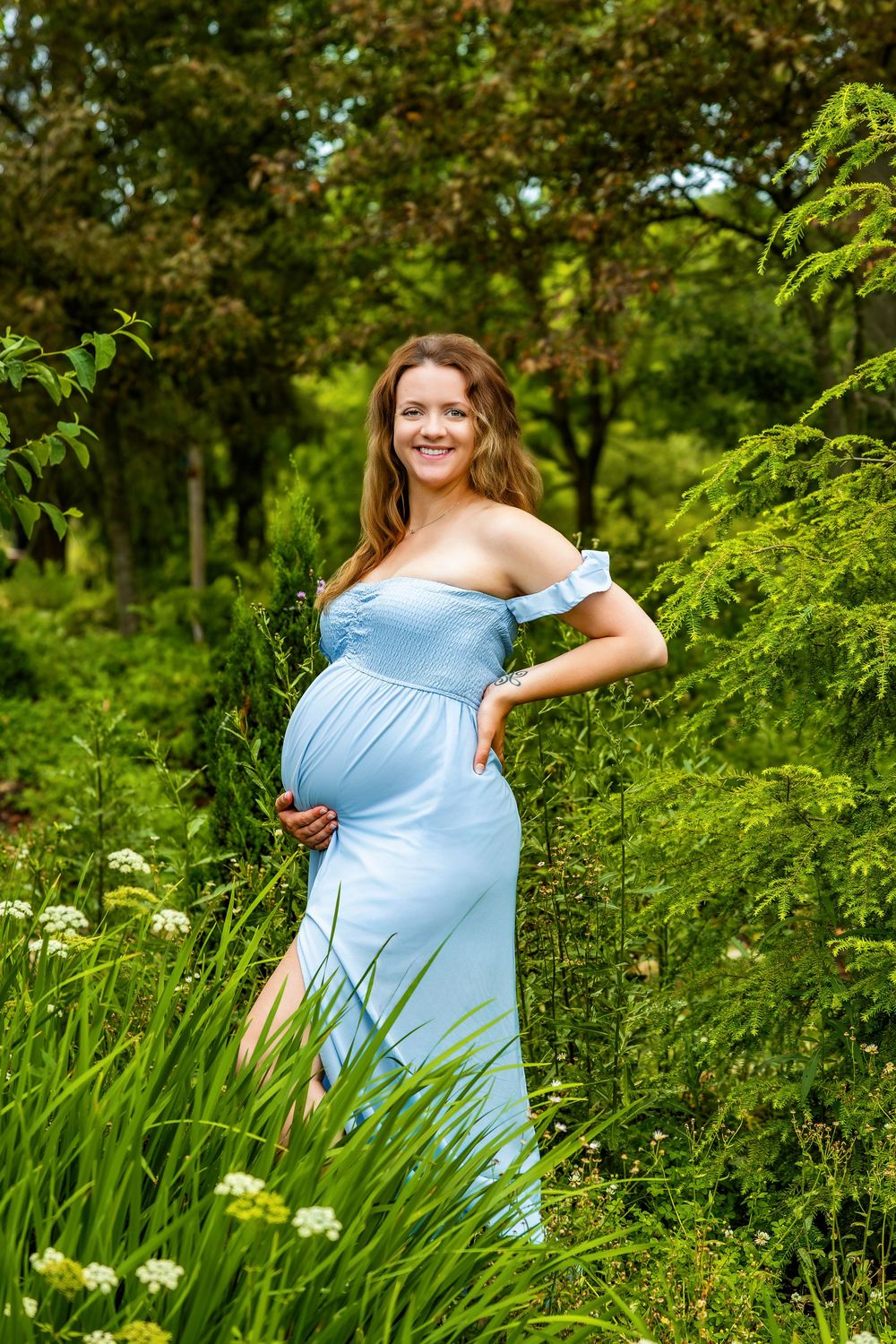 A maternity portrait features an expecting mother in a flowing blue dress amid vibrant garden greenery.