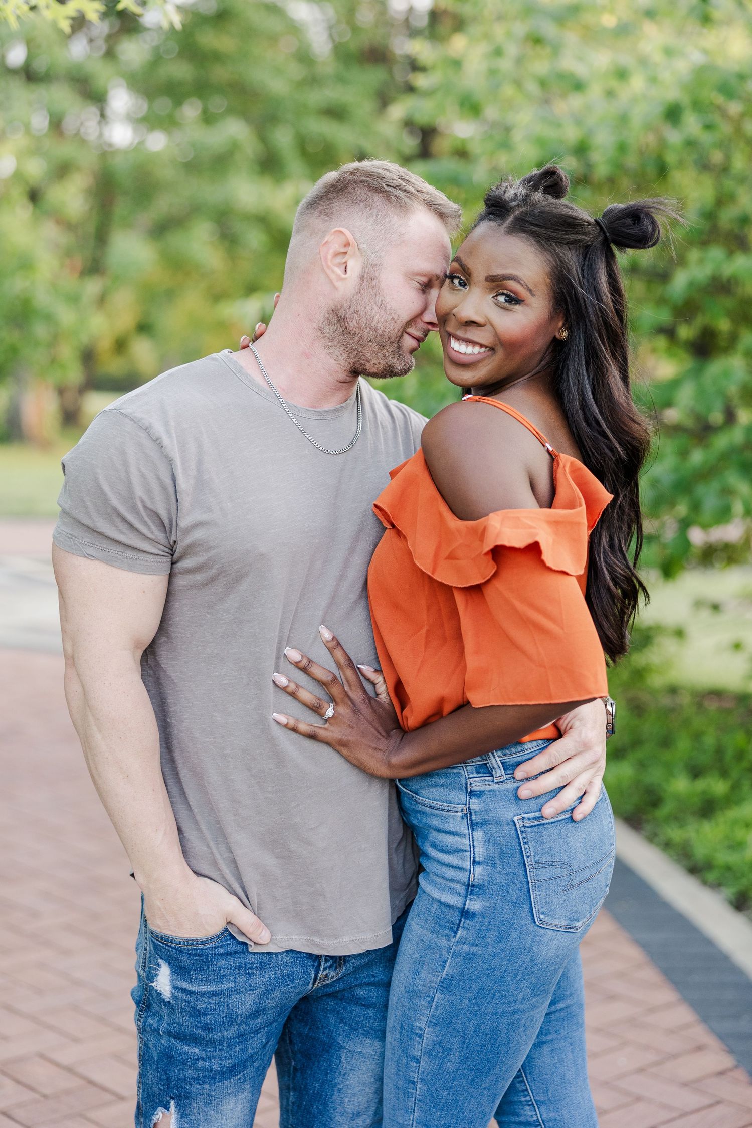 Happy couple embraces outdoors on brick path with green foliage background, wearing casual summer outfits.