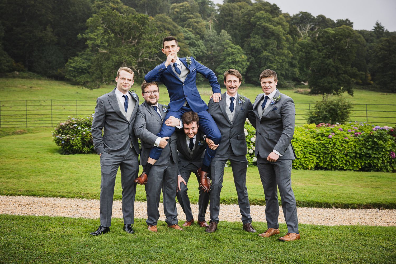 A group of groomsmen in gray suits pose playfully on a lush green lawn at an outdoor wedding venue.