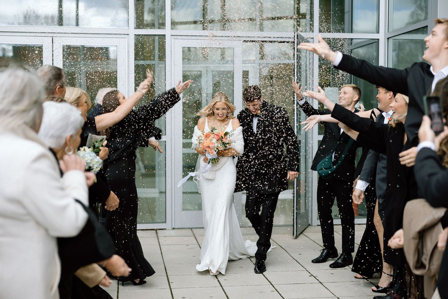 Wedding guests throw confetti over couple exiting glass doors while celebrating on outdoor terrace.