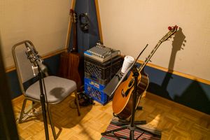 Recording studio setup with acoustic guitar on stand, microphones, and audio equipment on wooden parquet floor.