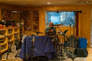 Professional recording studio setup with drums, microphones, and isolation booth visible through glass window.