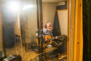 Musician plays acoustic guitar in a professional recording booth behind glass panels.