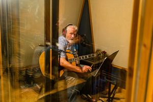 Musician playing acoustic guitar while reading sheet music in golden-lit recording studio booth.