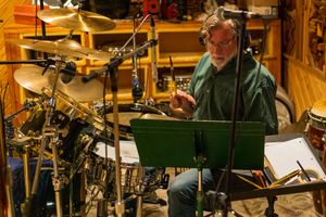 Drummer performs at a music stand surrounded by cymbals and percussion instruments in a wooden studio setting.