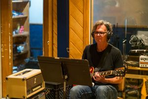 Musician wearing glasses plays electric guitar while seated in a recording studio with amplifier and equipment.