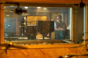A recording studio viewed through a control room window showing someone behind glass with warm lighting.