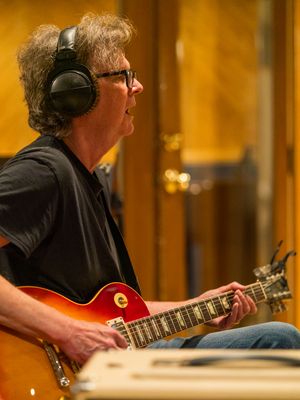 Musician wearing headphones plays red electric guitar in recording studio with warm lighting.