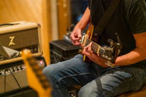 Close-up of guitarist playing electric guitar in recording studio with amplifier visible.