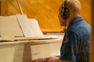 A musician playing a white piano while wearing headphones in a warm-lit studio.