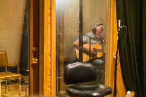 Acoustic guitarist performing in soundproof recording booth with glass partition.