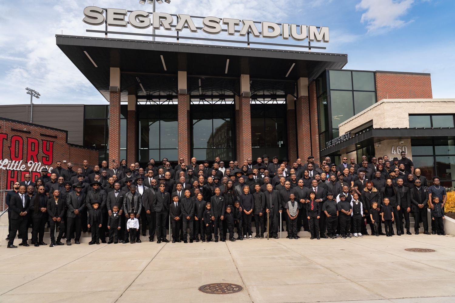 Large group of graduating students in black caps and gowns stand outside a sports stadium building for a graduation photo.