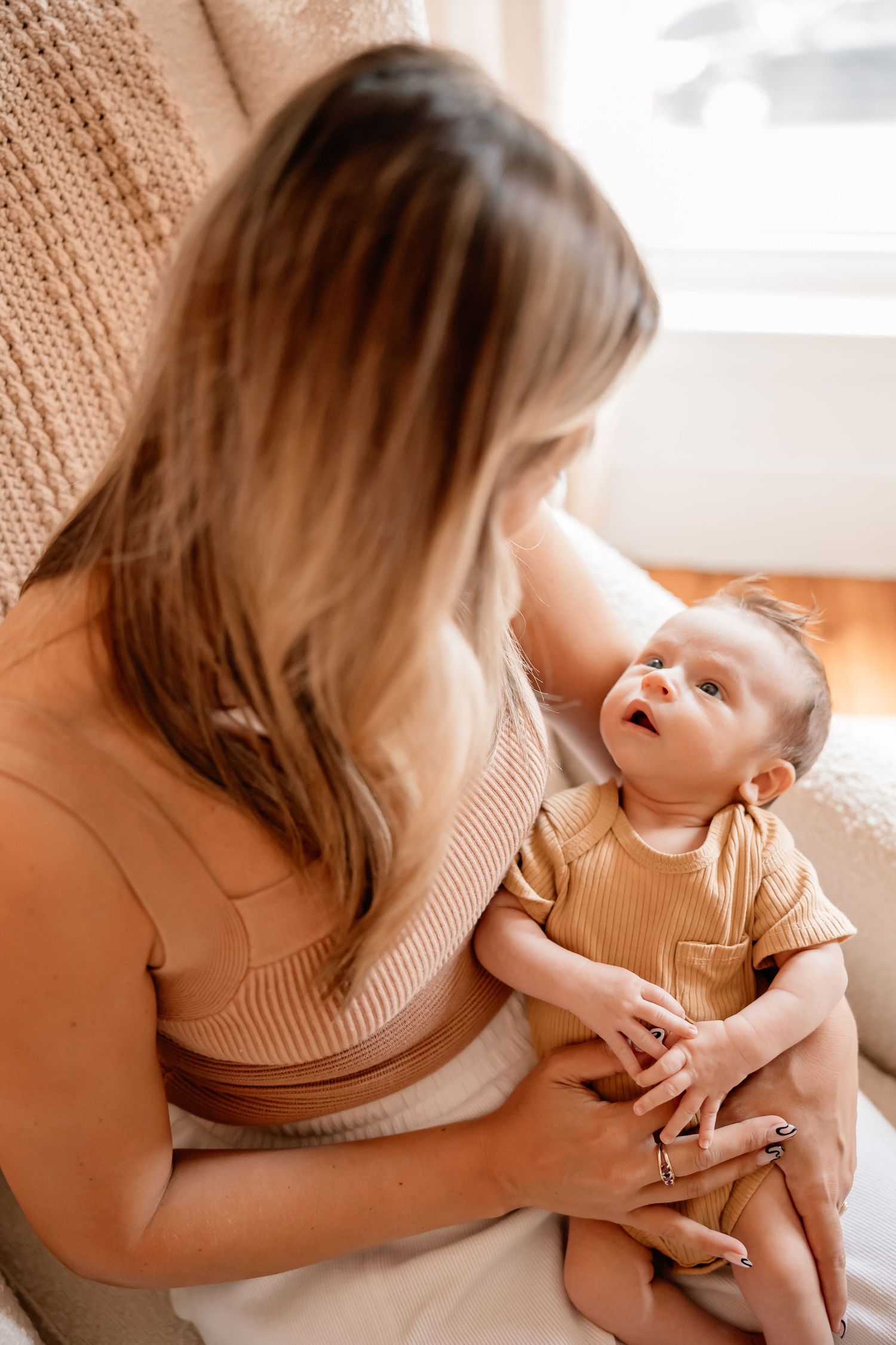 A happy baby laughs while being held in gentle embrace, both wearing cozy beige knit sweaters in warm indoor lighting.