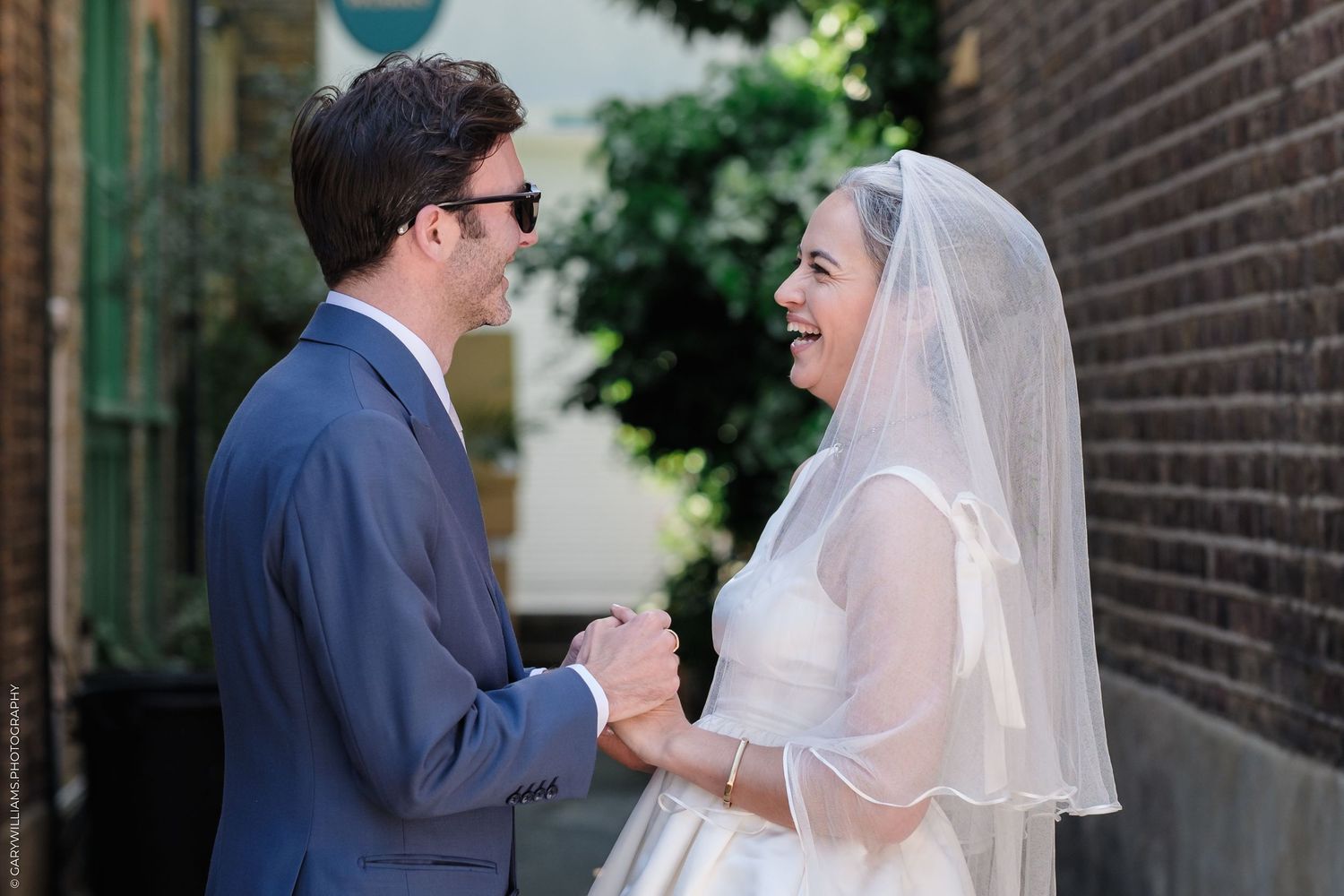 A bride in a white wedding dress and veil shares a joyful moment with her groom in a navy suit by a brick wall.