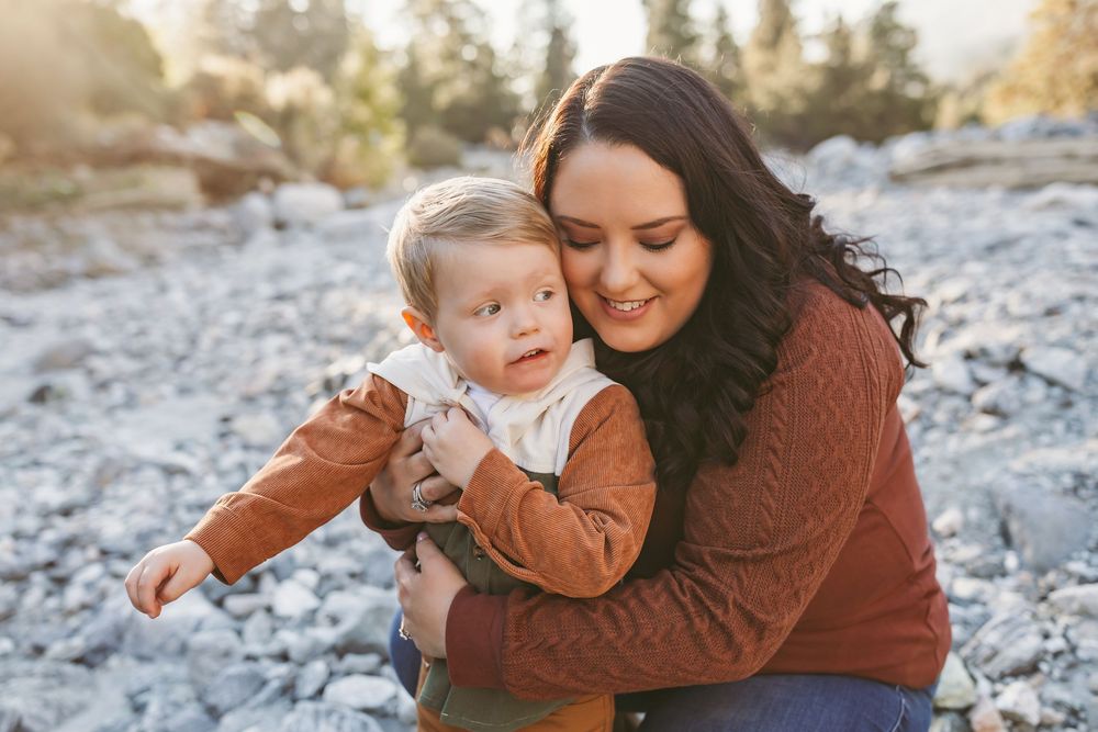 Woman in brown sweater holding small child wearing orange outfit sits together on rocky terrain during sunset.