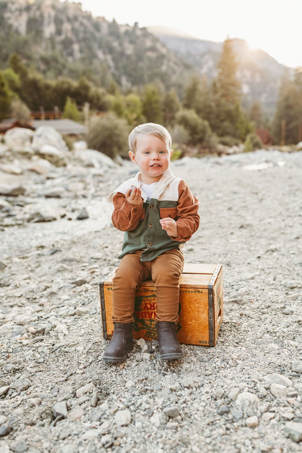 Toddler sitting on antique wooden crates in rocky mountain setting during golden hour sunset.