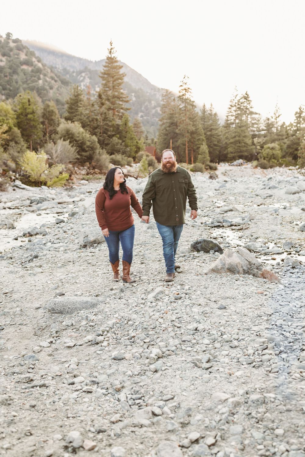 Two people walk hand in hand along a rocky mountain trail surrounded by evergreen trees in golden sunlight.