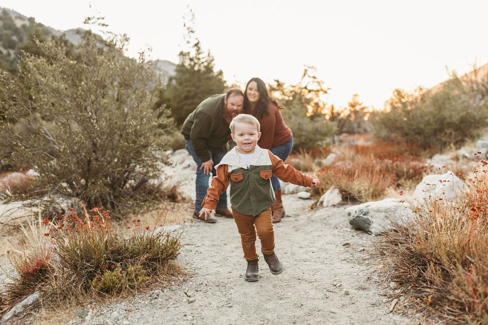 Young child runs joyfully down a nature trail path at sunset with family following behind in warm autumn lighting.