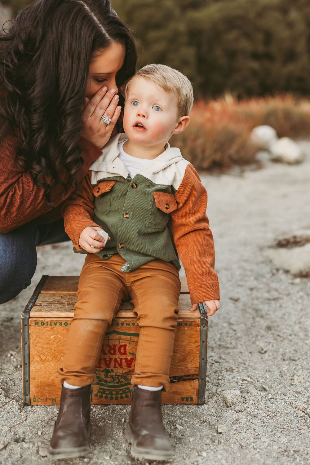 A toddler in olive green overalls and brown boots sits on a rustic wooden crate in a dreamy outdoor setting.