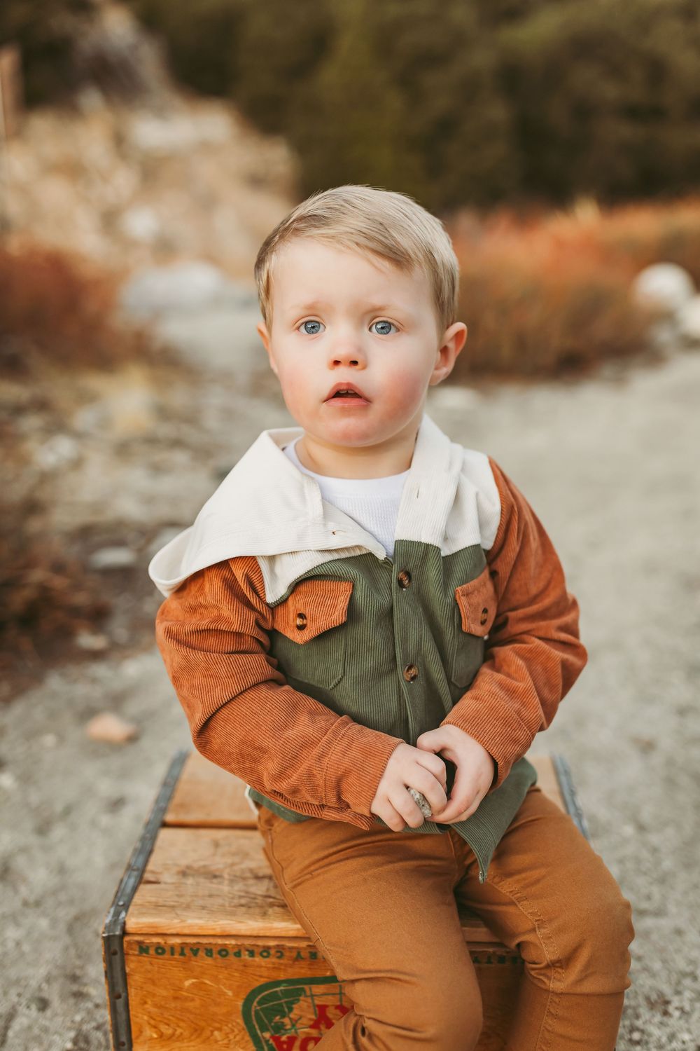 Small child wearing a colorblock jacket in rust and olive sits on vintage wooden crate in outdoor autumn setting.
