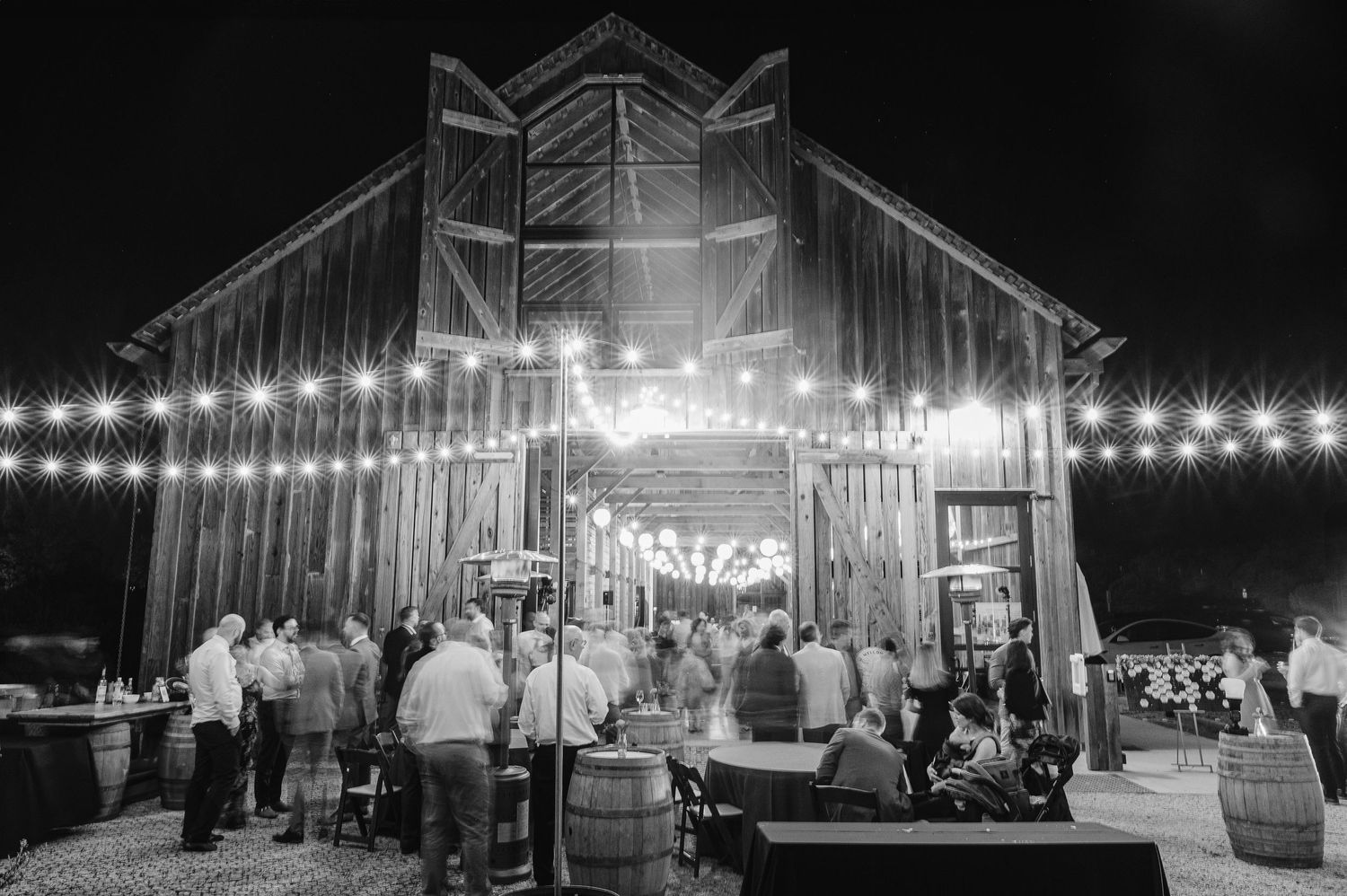A rustic barn venue illuminated by string lights at night with guests gathering outside for a celebration event.