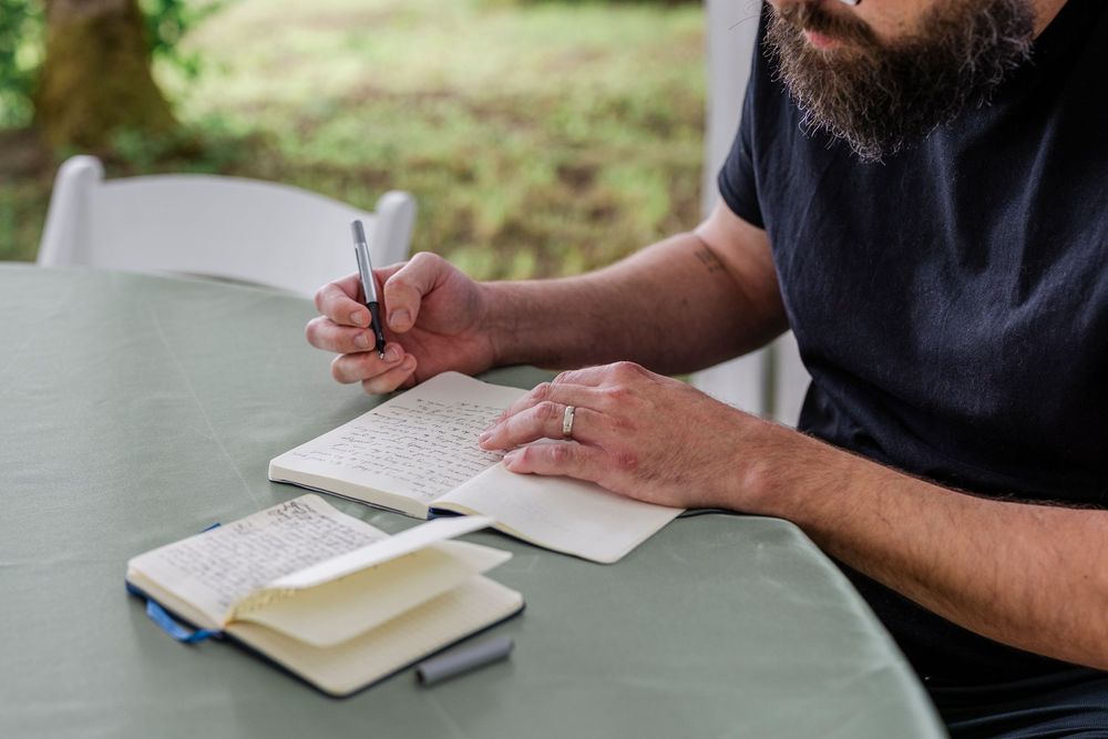 Person in navy shirt writes in notebook while sitting at outdoor table with books nearby.