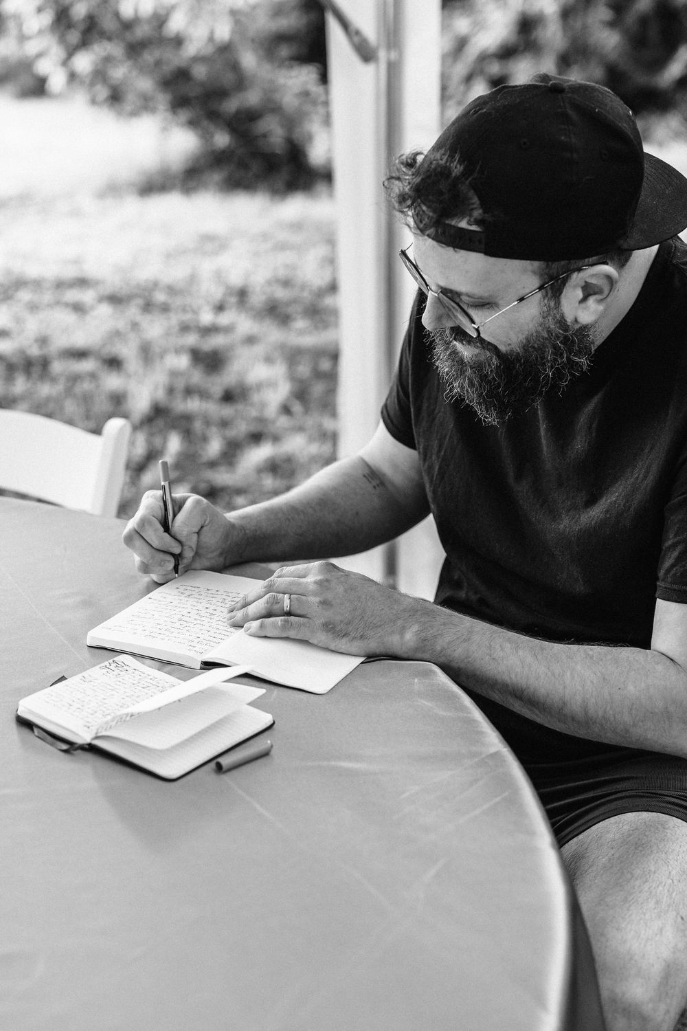 A bearded man in a baseball cap writes in a notebook while sitting at an outdoor table with books beside him.